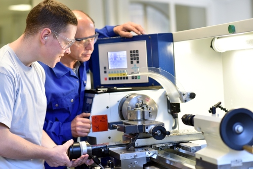 middle aged instructor wearing safety glasses instructs a young man, also wearing safety glasses, on how to operate a CNC machine. 